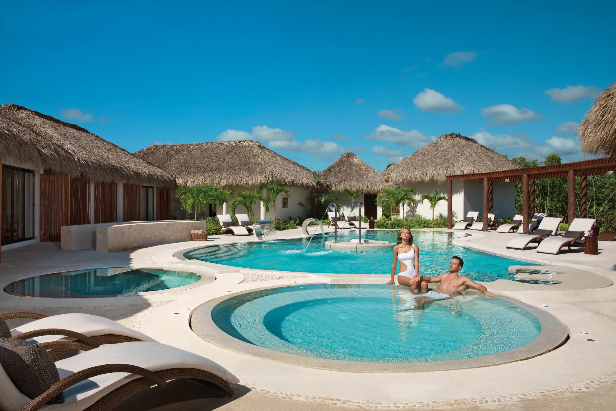 Guests relaxing in the outdoor hydrotherapy circuit pool at Secrets Cap Cana Resort & Spa, surrounded by tropical thatched-roof spa villas in Punta Cana.